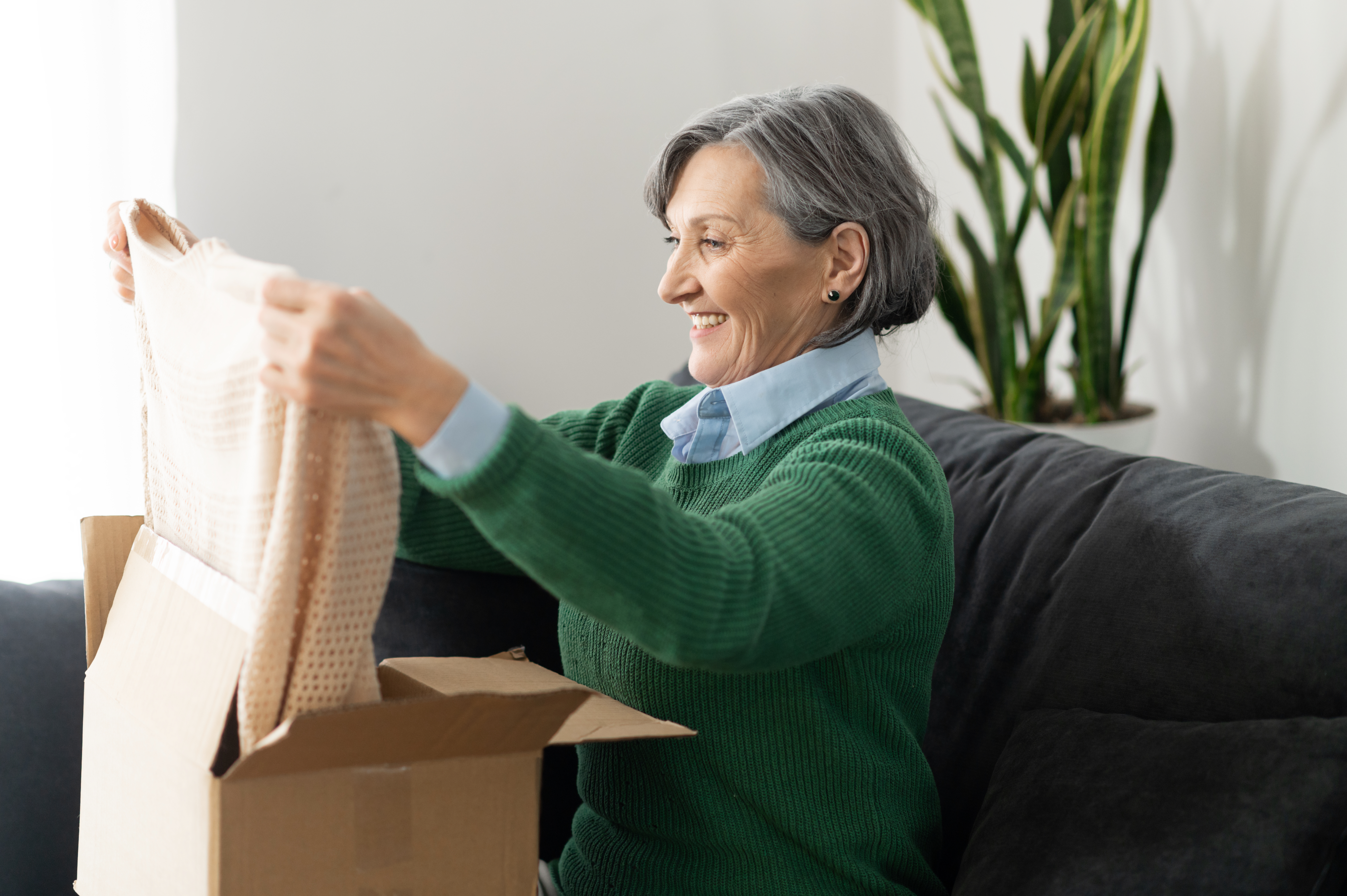 A woman sitting on a sofa and smiling as she unpacks a beige jumper. 